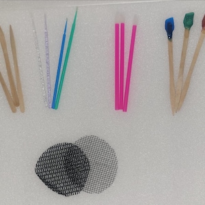 Op de afbeelding: Assortiment van cosmetische applicators en gereedschappen. Inclusief houten applicators, microborstels in helder en gekleurd plastic, roze lipwands en applicators met blauwe, groene en rode tips. Twee zwarte mesh cirkels zijn ook aanwezig.