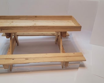 May include: A small, unfinished wooden picnic table with attached benches. The table has a rectangular top with a raised edge, and the benches are made of parallel wooden planks. The table is constructed from light-colored wood.
