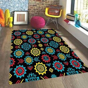 Puede incluir: Una alfombra negra con un patrón vibrante y repetitivo de formas estilizadas similares al sol en amarillo, rojo y turquesa. El diseño de la alfombra incluye flechas y elementos geométricos, creando una pieza audaz para un espacio habitable.