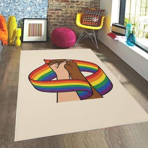 Puede incluir: Una alfombra rectangular con fondo crema presenta un gráfico de dos manos entrelazadas, rodeadas por una cinta de arcoíris. La alfombra está colocada sobre un suelo de madera, con un ambiente colorido en el fondo.