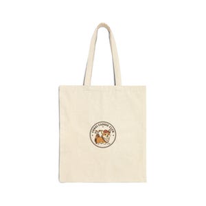 May include: A cream-coloured tote bag featuring a circular graphic of a corgi dog carrying golf clubs, with the text "Corgi Caddie Club". The bag has two long handles and is made of a sturdy fabric.