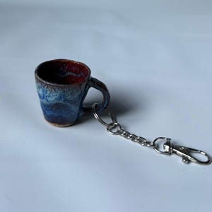 Op de afbeelding: Een miniatuur mok sleutelhanger van keramiek met een reactief glazuur in diepblauwe en bruine tinten. Het kleine kopje heeft een afgerond oortje bevestigd aan een zilverkleurige metalen ring, een kettinkje en een karabijnsluiting.