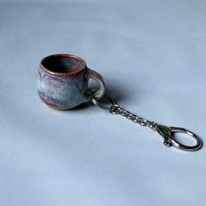Op de afbeelding: Een sleutelhanger met een miniatuur keramische mok, met een ronde vorm en een reactief glazuur in tinten blauw, paars en aards bruin. De mok is bevestigd aan een zilverkleurige metalen ketting met een ring en een karabijnsluiting.