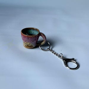 Op de afbeelding: Een miniatuur mok sleutelhanger van keramiek met een reactief glazuur in de kleuren donkerroze, beige en turquoise. Het kleine kopje heeft een gebogen oortje en is bevestigd aan een zilverkleurige metalen ring en een karabijnsluiting.