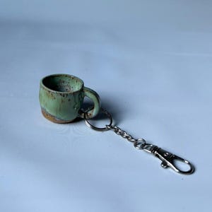 Op de afbeelding: Een miniatuur keramische mok sleutelhanger met een gespikkeld lichtgroen glazuur en bruine accenten aan de basis. De mok is bevestigd aan een zilverkleurige metalen ring, een korte ketting en een karabijnhaak voor een veilige bevestiging.