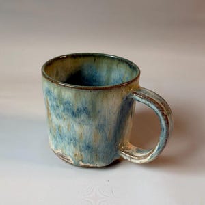 May include: A ceramic mug featuring a reactive glaze with blended shades of blue, cream, and earthy brown. The cylindrical vessel has a thick, curved handle and a slightly uneven, textured base with visible clay tones at the bottom rim.