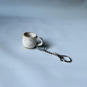 Op de afbeelding: Een miniatuur mok sleutelhanger van keramiek met een gespikkelde witte en beige afwerking. Het kleine, ronde kopje heeft een oortje dat is bevestigd aan een zilverkleurige metalen ring, een kort kettinkje en een karabijnsluiting.
