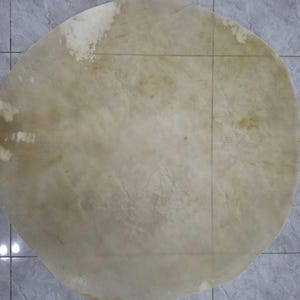 May include: A large, circular, off-white drumhead with visible grain and texture. The drumhead has a natural, organic appearance with subtle variations in color and tone. It is likely made of animal hide.