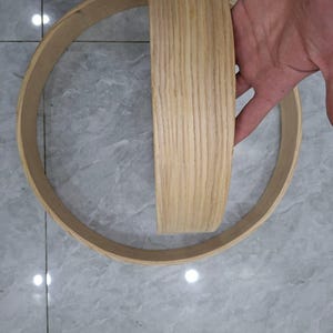 May include: A light-colored wooden hoop with a flat, wide band. The wood grain is visible on the surface. The hoop is held by a hand, showing the scale of the object. The item appears to be a component of a larger structure.