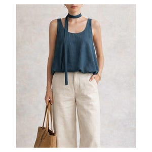May include: A teal tank top with a matching neck tie and cream-colored linen pants. The top has a scoop neckline and a relaxed fit. The pants are wide-legged and high-waisted. A woven tote bag is held in the hand.