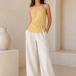 May include: A mustard yellow sleeveless halter top with a high neckline and a side slit, paired with off-white wide-leg trousers featuring a pleated front and a relaxed fit. The outfit is completed with simple tan sandals and a delicate gold necklace.