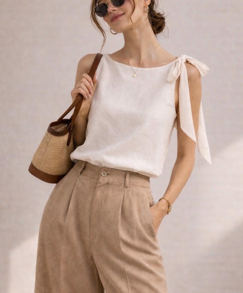 May include: A sleeveless off-white top featuring shoulder tie straps, paired with high-waisted tan trousers with front pleats and a button closure. A woven straw handbag with brown leather trim is carried at the side of the outfit.