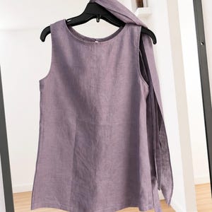 May include: A sleeveless lavender top with a round neckline and a long, integrated fabric tie draped over one shoulder. The garment is made of a textured fabric and hangs from a black plastic hanger against a dark-framed mirror.