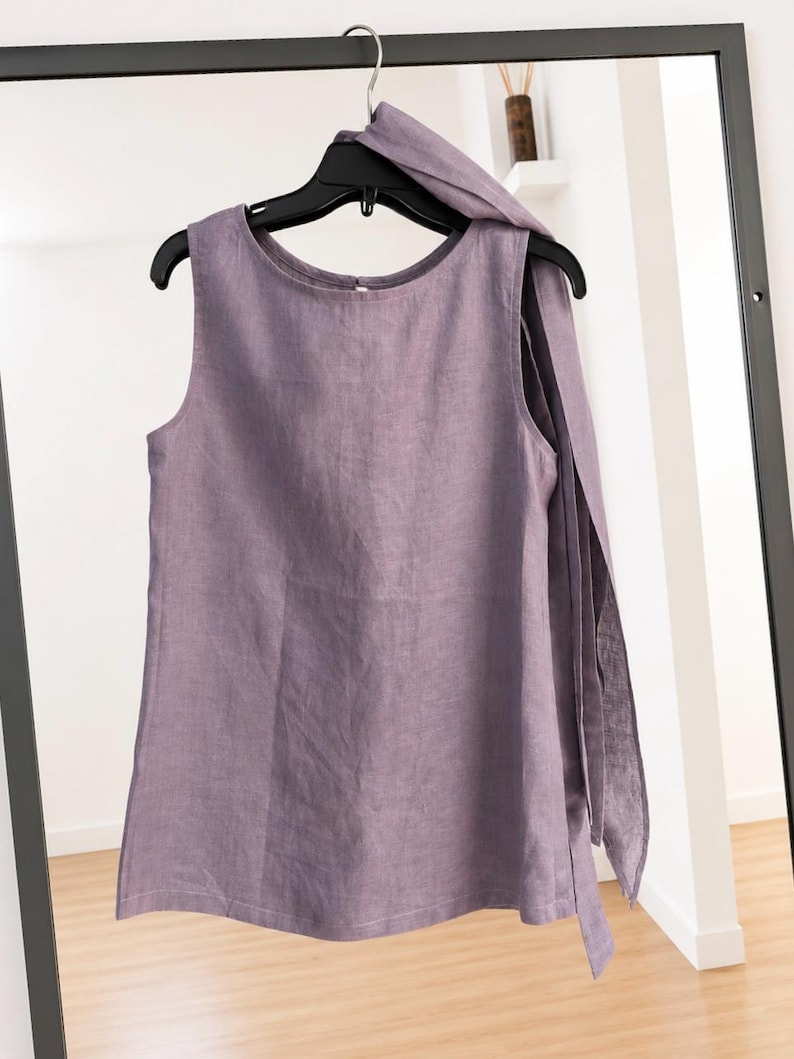May include: A sleeveless lavender top with a round neckline and a long, integrated fabric tie draped over one shoulder. The garment is made of a textured fabric and hangs from a black plastic hanger against a dark-framed mirror.