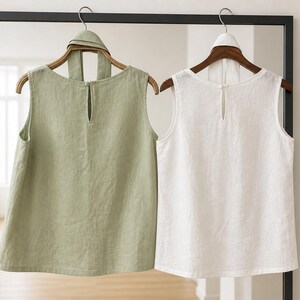 May include: Two sleeveless tops hanging on wooden hangers, one in sage green and one in white. Each top features a round neckline, a keyhole opening at the center back with a button closure, and a matching fabric tie detail at the collar.