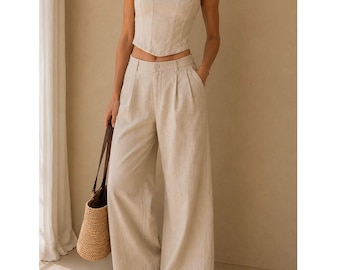Puede incluir: Un top estilo corsé de color beige con tirantes finos y escote festoneado, combinado con pantalones anchos de cintura alta a juego con pliegues delanteros. Un bolso de paja trenzada con asas de cuero marrón y sandalias de cuero marrón completan el conjunto.