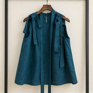 May include: A sleeveless teal top featuring a round neckline and shoulder straps that tie into bows. The garment has a loose, flared silhouette and two long, narrow fabric strips hanging down from the front neckline, all made of a textured fabric.