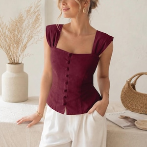 May include: A sleeveless deep burgundy top featuring a square neckline, wide shoulder straps, and a vertical row of matching fabric-covered buttons down the center front. The bodice is fitted with a straight hemline, paired with white trousers.