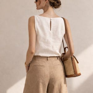 May include: A sleeveless white top with a small button closure at the back neckline, paired with tan wide-leg trousers. A structured straw bucket bag with brown leather trim and a long shoulder strap hangs from the shoulder.