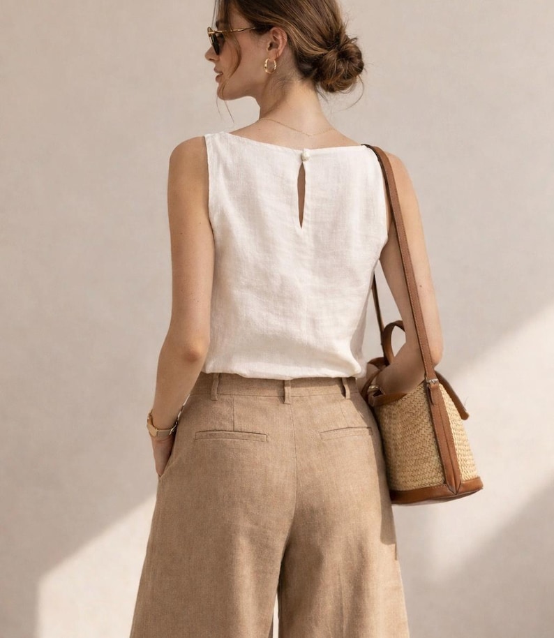 May include: A sleeveless white top with a small button closure at the back neckline, paired with tan wide-leg trousers. A structured straw bucket bag with brown leather trim and a long shoulder strap hangs from the shoulder.