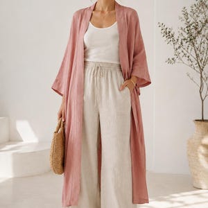 May include: A dusty rose open-front duster cardigan layered over a white scoop-neck tank top and beige wide-leg drawstring trousers. The outfit is paired with tan woven slide sandals and a small round straw handbag, creating a coordinated ensemble.