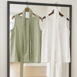 May include: Two sleeveless tops hanging on wooden hangers, one in sage green and one in white. Each top features a round neckline and a long, matching fabric tie attached at the collar that hangs down the front of the garment.