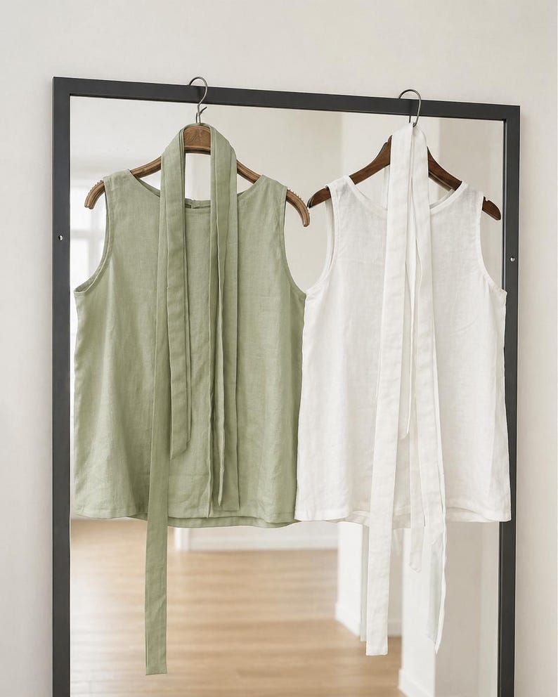 May include: Two sleeveless tops hanging on wooden hangers, one in sage green and one in white. Each top features a round neckline and a long, matching fabric tie attached at the collar that hangs down the front of the garment.