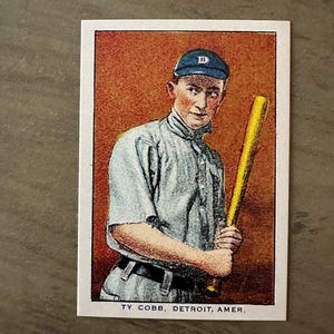 Può includere: Carta da baseball vintage con Ty Cobb di Detroit, che tiene una mazza gialla. Indossa un berretto blu con una "D" bianca e un'uniforme azzurra. La carta ha uno sfondo color seppia e il testo "TY COBB, DETROIT, AMER."