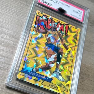 Op de afbeelding: Een beoordeelde Panini Kaboom! basketbalkaart met Kobe Bryant in een Lakers-uniform. De kaart heeft een geel en blauw ontwerp met het woord "KABOOM!" en het Lakers-logo. De kaart is ingesloten in een beschermende plastic houder.