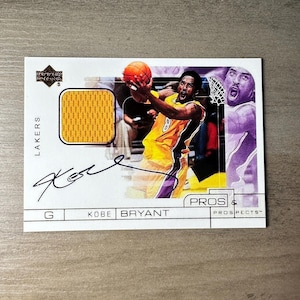 Könnte beinhalten: Eine Sammler-Basketballkarte von Upper Deck mit Kobe Bryant im Lakers-Trikot. Die Karte hat ein Trikotmuster und eine Unterschrift. Die Karte zeigt ein Foto von Kobe Bryant in Aktion mit dem Text "PROS & PROSPECTS".