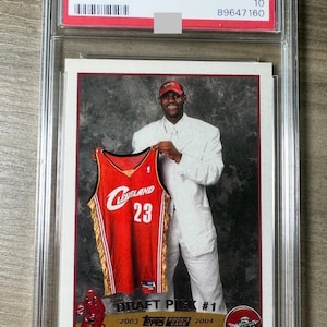 May include: A 2003 Topps LeBron James rookie card, graded GEM MT 10, encased in a protective holder. The card features James holding a Cleveland Cavaliers jersey with the number 23. The card is labeled "Draft Pick #1".