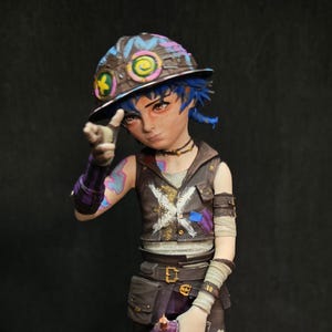 Puede incluir: Figura detallada de un personaje con cabello azul, que lleva un chaleco marrón, leggings morados y botas. La figura sostiene un arma azul y plateada. La base presenta texto estilo grafiti, incluyendo la palabra "Jinx" y "HAHA". También hay una linterna azul.