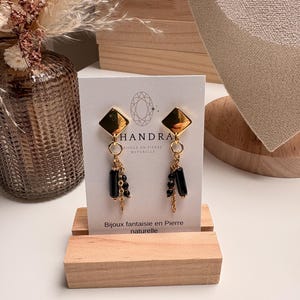 May include: A pair of gold-colored, diamond-shaped earrings with black rectangular beads and delicate chain accents. The earrings are displayed on a white card with the brand name "HANDRA" and the text "Bijoux fantaisie en Pierre naturelle".