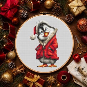 Op de afbeelding: Een kruissteek borduurproject in een ronde houten borduurring met een cartoonpinguïn die een rode kerstmuts en een rode jas met een beige bontkraag draagt. De pinguïn heeft zwart-witte veren, oranje voeten en een snavel.