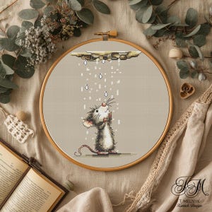May include: A cross-stitch embroidery hoop showing a cartoon rat looking up at raindrops. The rat is gray and white, with a pink nose and a long tail. The background is a light beige fabric, and the wooden hoop is visible.