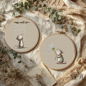 Könnte beinhalten: Zwei Kreuzstich-Stickrahmen mit Mäuse-Illustrationen. Einer zeigt eine Maus unter einer Regenwolke, der andere mit einem Löwenzahn. Die Rahmen sind vor einem neutralen Stoffhintergrund mit Holzrahmen angeordnet.