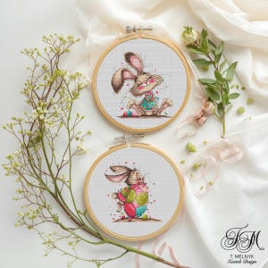 May include: Two cross-stitch hoops featuring Easter bunny designs. One shows a bunny with a colorful bib and eggs, the other with a bunny hugging a decorated egg. The hoops are set against a white background with floral accents.
