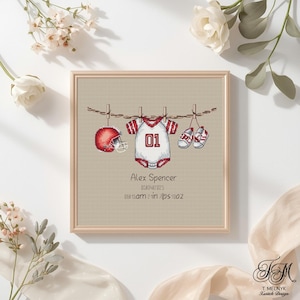 May include: Cross-stitch artwork in a wooden frame featuring a baby football theme. Includes a red helmet, a onesie with the number 01, and baby shoes. The text reads "Alex Spencer" and birth details.