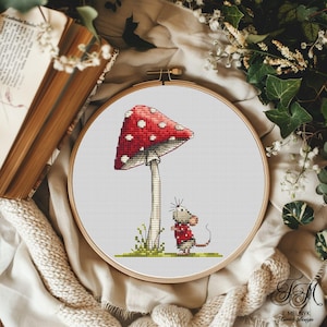 May include: A cross-stitch embroidery in a wooden hoop features a red mushroom with white spots and a small mouse wearing a red sweater. The mouse stands near the mushroom. The embroidery is set against a neutral background.