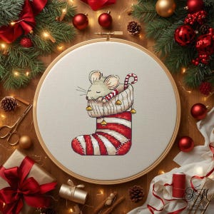May include: A cross-stitch embroidery in a wooden hoop featuring a light-coloured mouse tucked inside a red and white striped Christmas stocking. The stocking is decorated with small gold bells and a candy cane handle, set against a white fabric background.