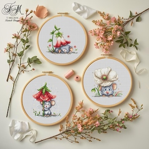 Könnte beinhalten: Drei Kreuzstich-Stickrahmen mit entzückenden Maus-Designs unter Blumen. Die Rahmen sind vor einem Hintergrund aus Blumenarrangements und einer weißen Oberfläche angeordnet. Die Designs zeigen eine Maus unter einer roten Blume, einer weißen Blume und einer roten Glockenblume.