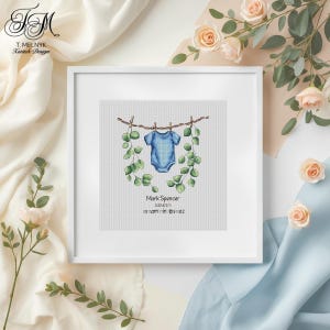 May include: A framed cross-stitch artwork featuring a blue baby onesie hanging on a clothesline, adorned with green foliage. The artwork includes the name "Mark Spencer", date, time, and weight in pounds and ounces.