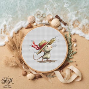 May include: Cross-stitch embroidery of a mouse wearing a straw hat with a red ribbon, set in a wooden hoop. The artwork is surrounded by seashells, scissors, and other beach-themed elements on a sandy surface.