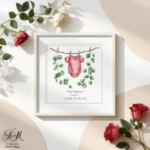 May include: A framed cross-stitch design featuring a pink baby onesie hanging on a clothesline with green foliage. The text "Anna Spencer" and other details are below the onesie. The frame is white.