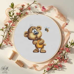 May include: A cross-stitch embroidery hoop showing a cartoon bear looking at a bee. The bear is brown with a yellow belly, and the bee is black and yellow. The embroidery is set against a white background.