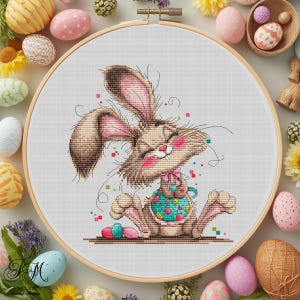 Puede incluir: Bordado de punto de cruz de un conejito sonriente en un aro de madera. El conejito es marrón con detalles rosas y un delantal turquesa, rodeado de huevos de Pascua y flores. Una encantadora decoración de Pascua hecha a mano.