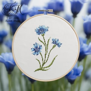 Può includere: Un ricamo a punto croce di fiordalisi blu su tessuto bianco, incorniciato in un telaio di legno. I fiori hanno petali blu dettagliati e steli verdi. Il testo "T. Melnyk Xstitch Design" è visibile.