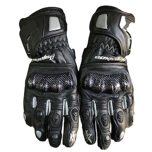 Könnte beinhalten: Ein Paar schwarze Motorradhandschuhe aus Leder mit silbernen Akzenten. Die Handschuhe verfügen über Knöchelschutz aus Kohlefaser, das Hayabusa-Branding und ein Kawasaki-Logo. Entwickelt für Schutz und Griffigkeit.