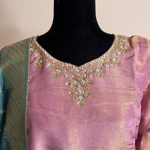 Pode incluir: Uma túnica de seda rosa com bordados dourados ao redor do decote e das mangas. O decote apresenta intrincados trabalhos de contas douradas e turquesas. Um lenço estampado em verde-azulado e dourado é drapeado sobre o ombro.
