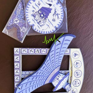 May include: Decorative blue and white ceramic coasters and a decorative frame. The coasters feature a circular design with a boat scene and a geometric pattern. The frame has a wing-like shape with a similar design.